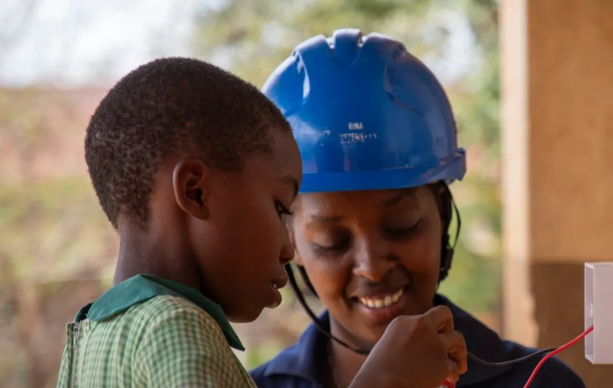 A schoolgirl and a woman electrician in a blue helmet fitting a light switch together at a Kenyan primary school